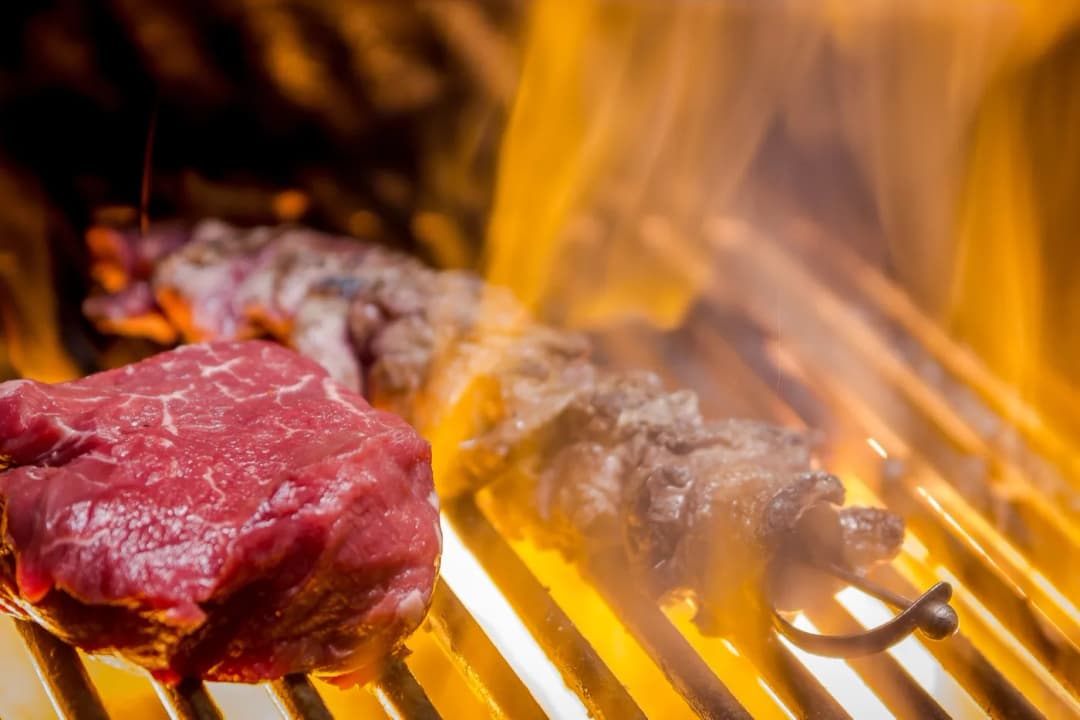 Raw steak and a meat skewer over open flames on the grill